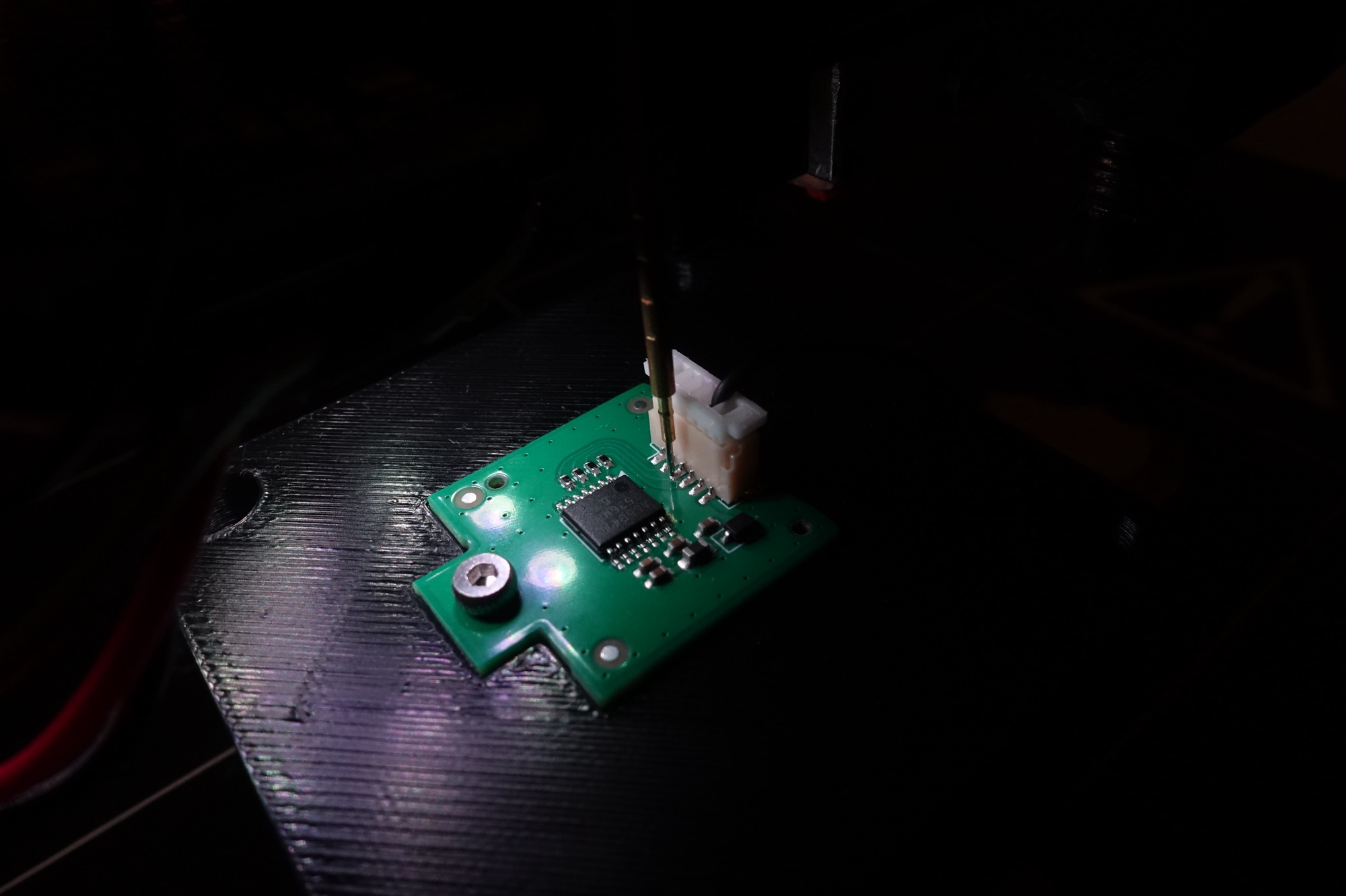 A gold pogo pin descending onto a small green PCB clamped on the machine bed, lit from one side in a dark room. The tip is landing on an exposed pad beside a white JST connector and an SOIC package.