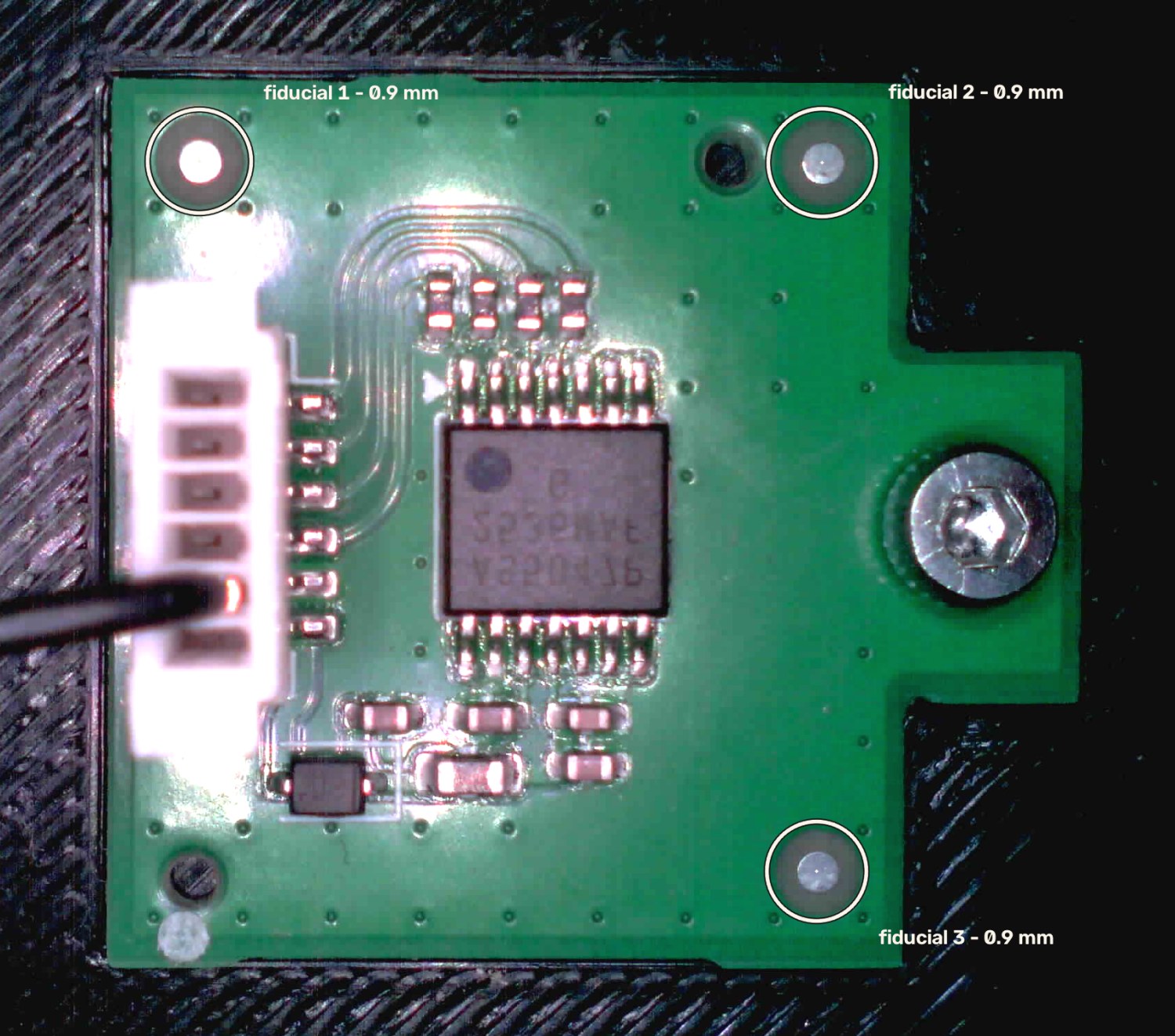 Overhead view of a small green PCB held in a black fixture, lit by a ring light. Three circular bare-copper pads near the board corners are ringed and labelled as fiducials, each 0.9 mm across. A white wire-to-board connector with a black cable sits at the left edge, an SOIC package at centre, and a steel mounting screw at the right.